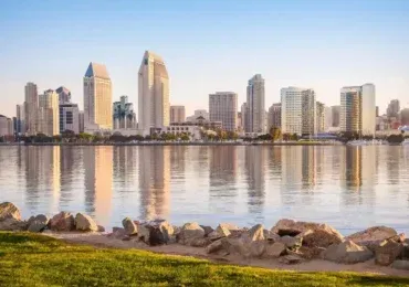 San Diego skyline reflecting on calm water, with a rocky shoreline in the foreground.