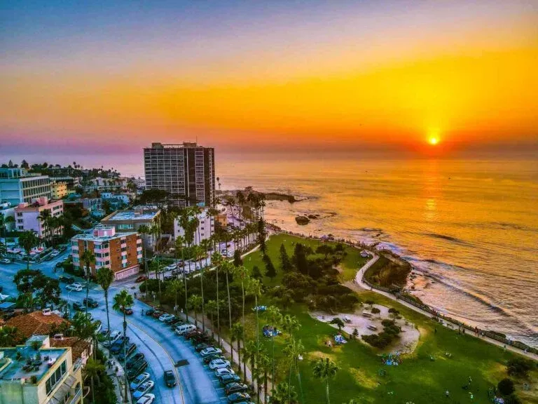 Sunset over the Pacific Ocean, illuminating a coastal city with buildings, palm trees, and a winding road.