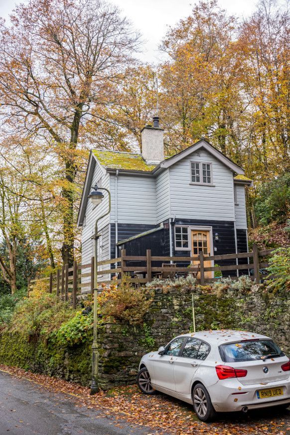 A white car is parked in front of a house in the woods. The house has a green mossy roof.