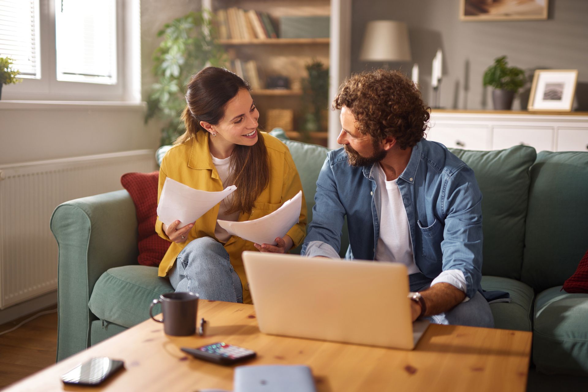 Couple on a couch reviewing papers with a laptop, smiling.