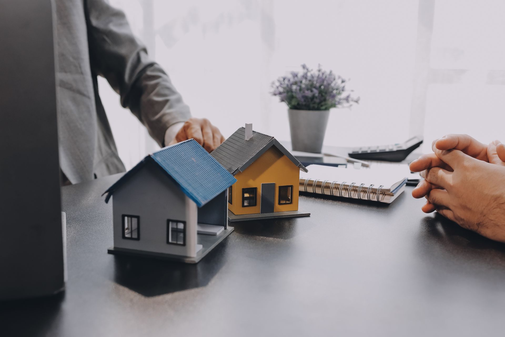 Two miniature houses on a desk, a person in a suit points, hands clasped next to a person.