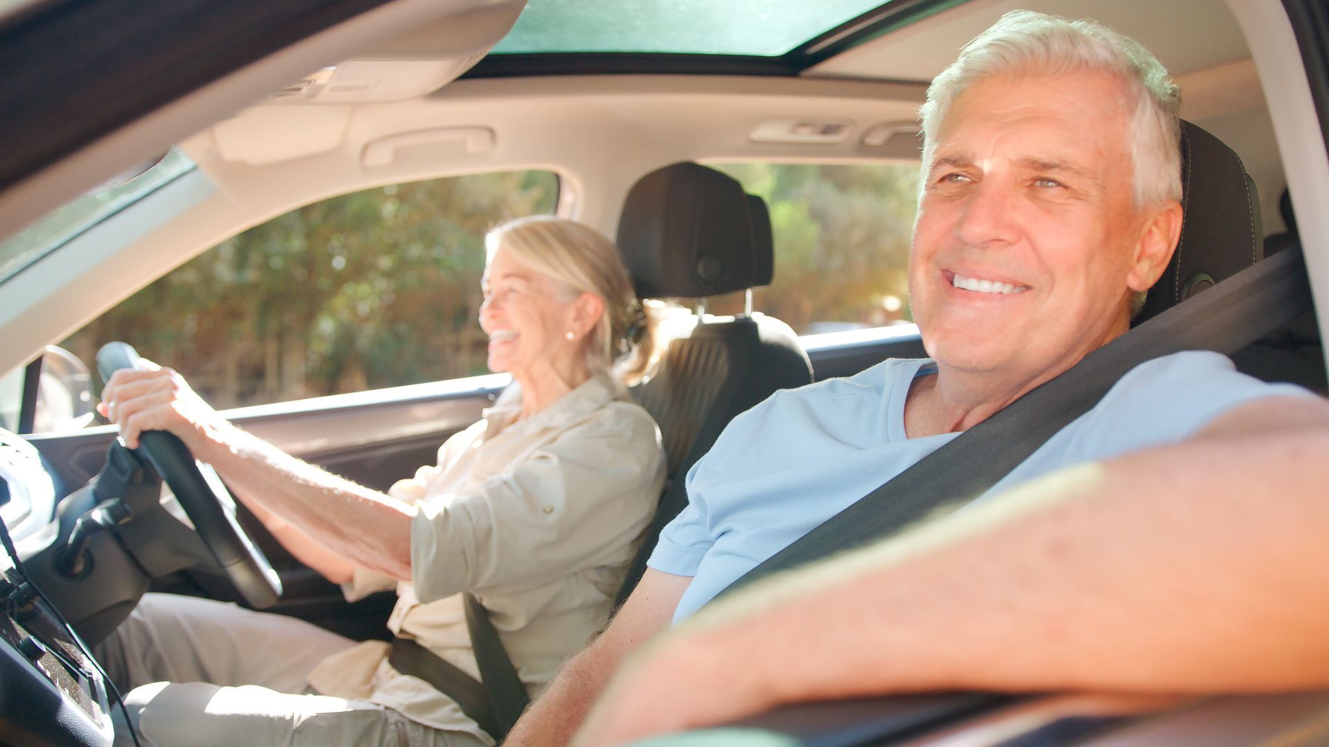Elderly couple in car, smiling. Woman driving, man passenger. Sunny outdoors.