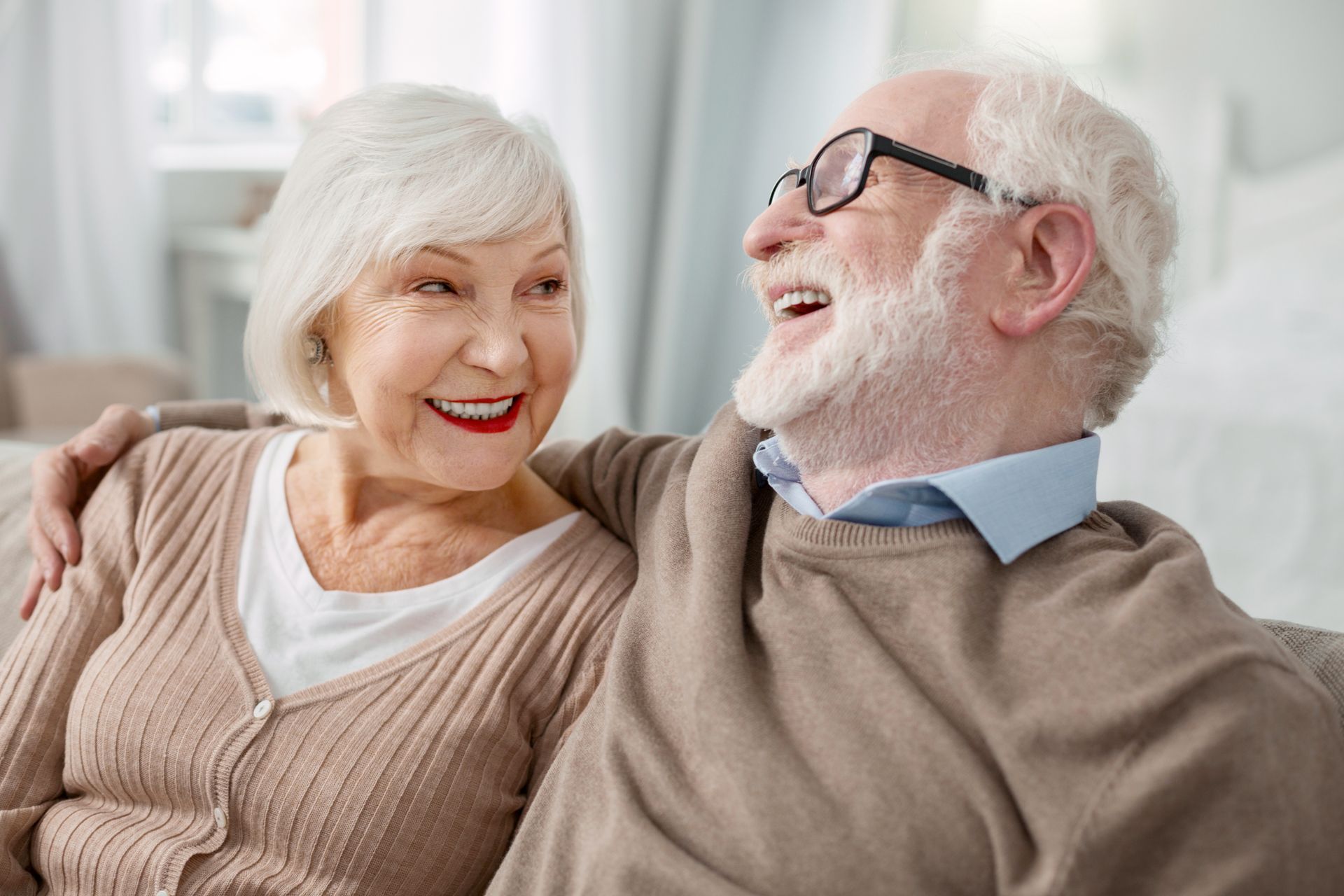 Smiling older couple sitting together, arm around shoulder. Bright indoor setting.