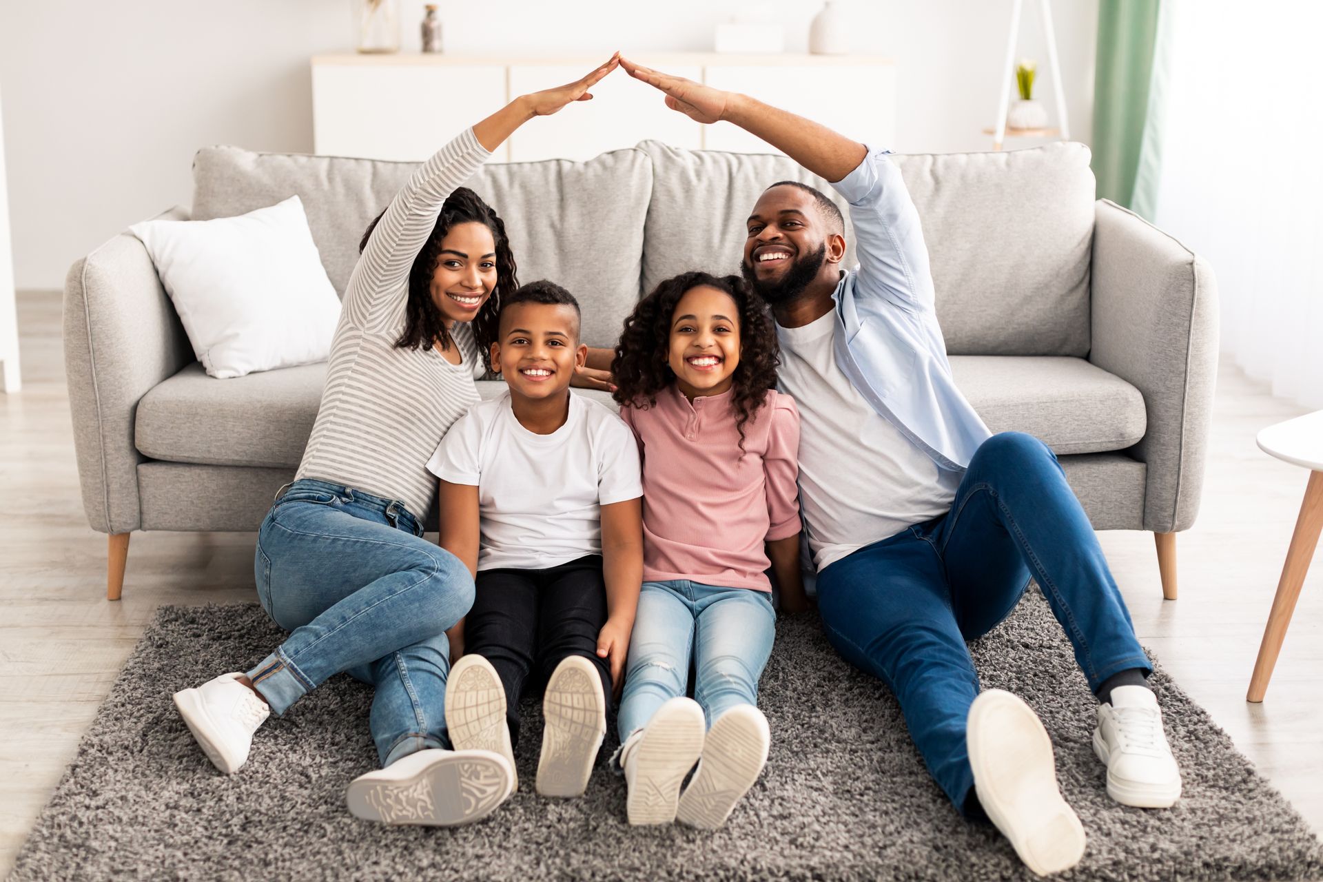 Family of four making a roof shape with arms while sitting on the floor in their living room.