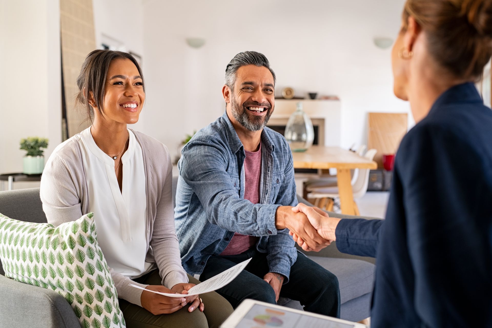 Couple smiles and shakes hands with a person in a living room; discussing paperwork.