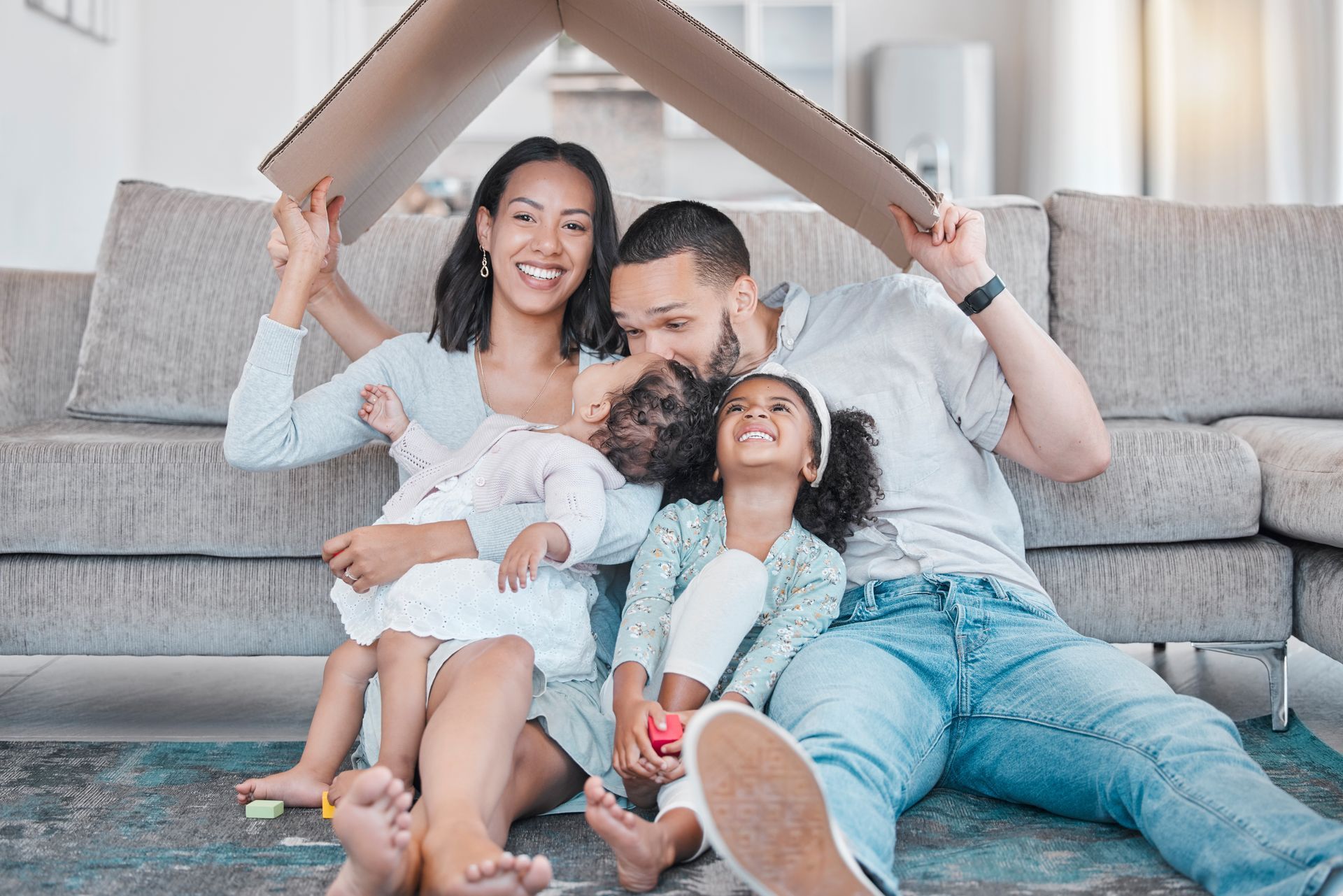 Family of four sitting on floor under cardboard house, smiling.