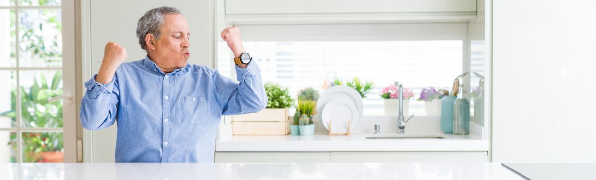 An elderly man is sitting at a table in a kitchen with his arms in the air.