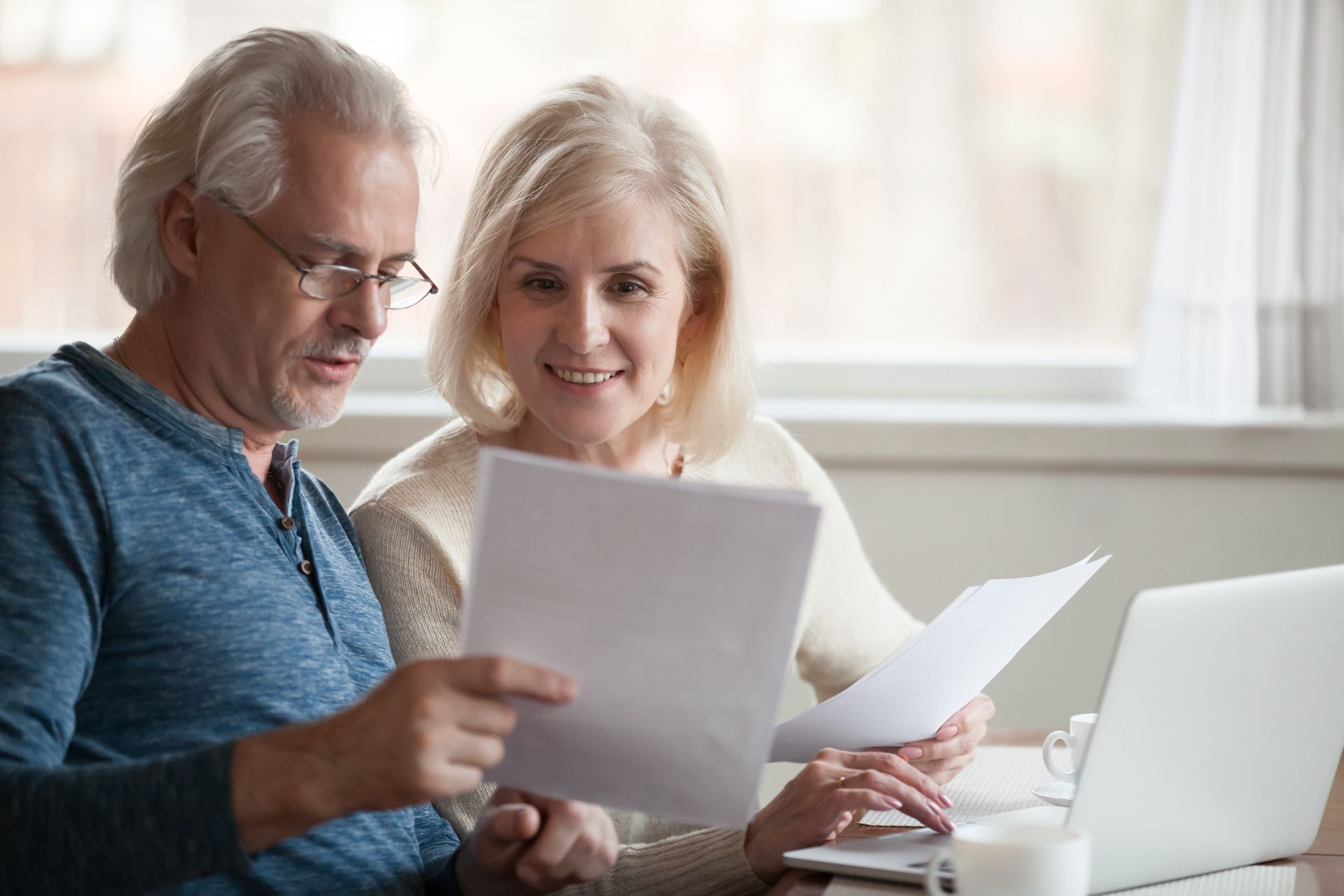 An elderly couple is sitting at a table looking at papers and a laptop.