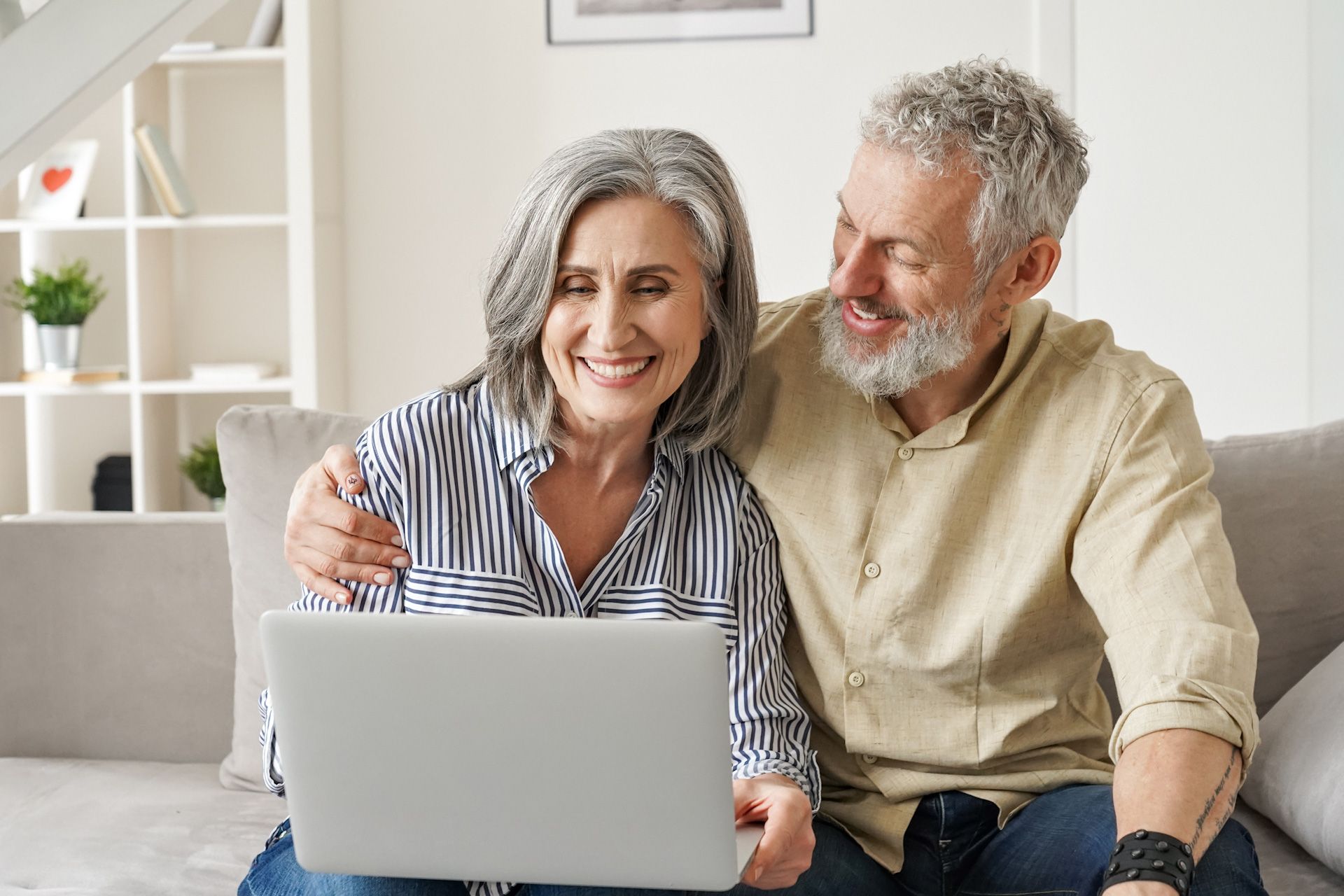 An elderly couple is sitting on a couch using a laptop computer.