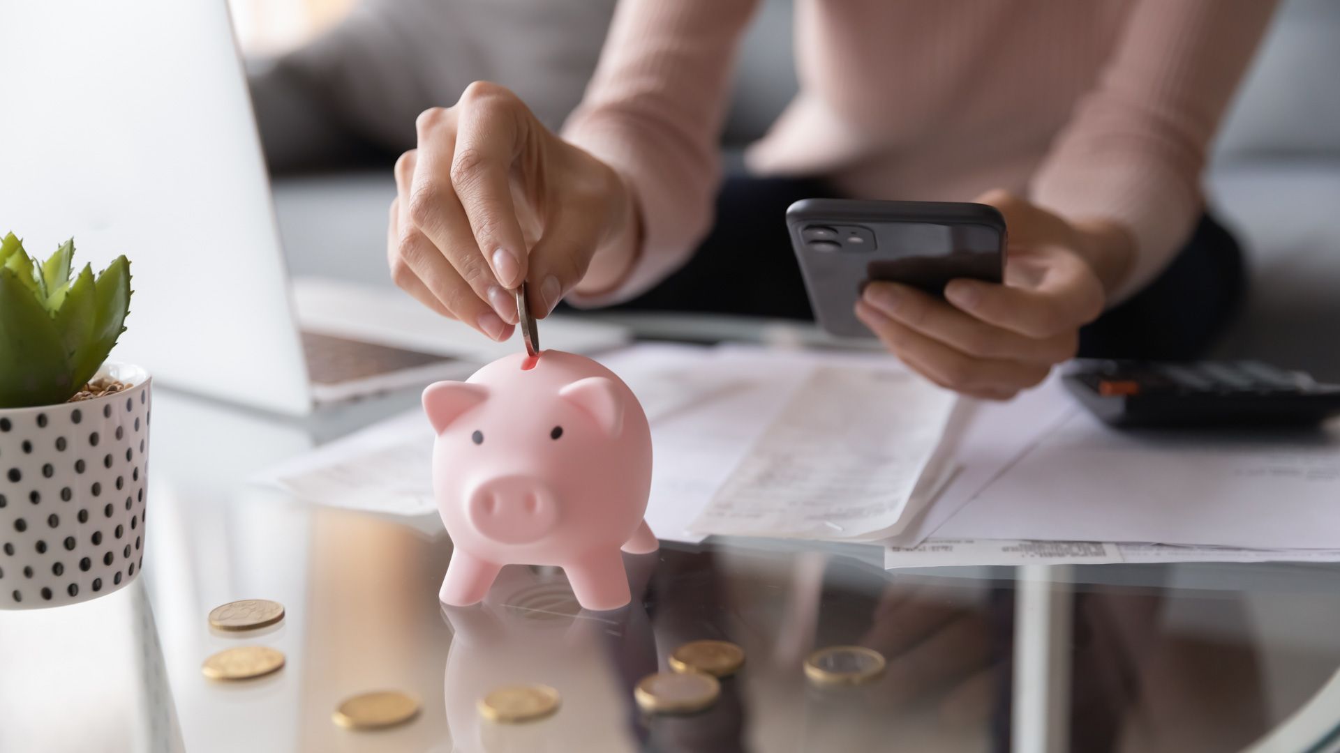 A woman is putting a coin into a piggy bank while using a cell phone.