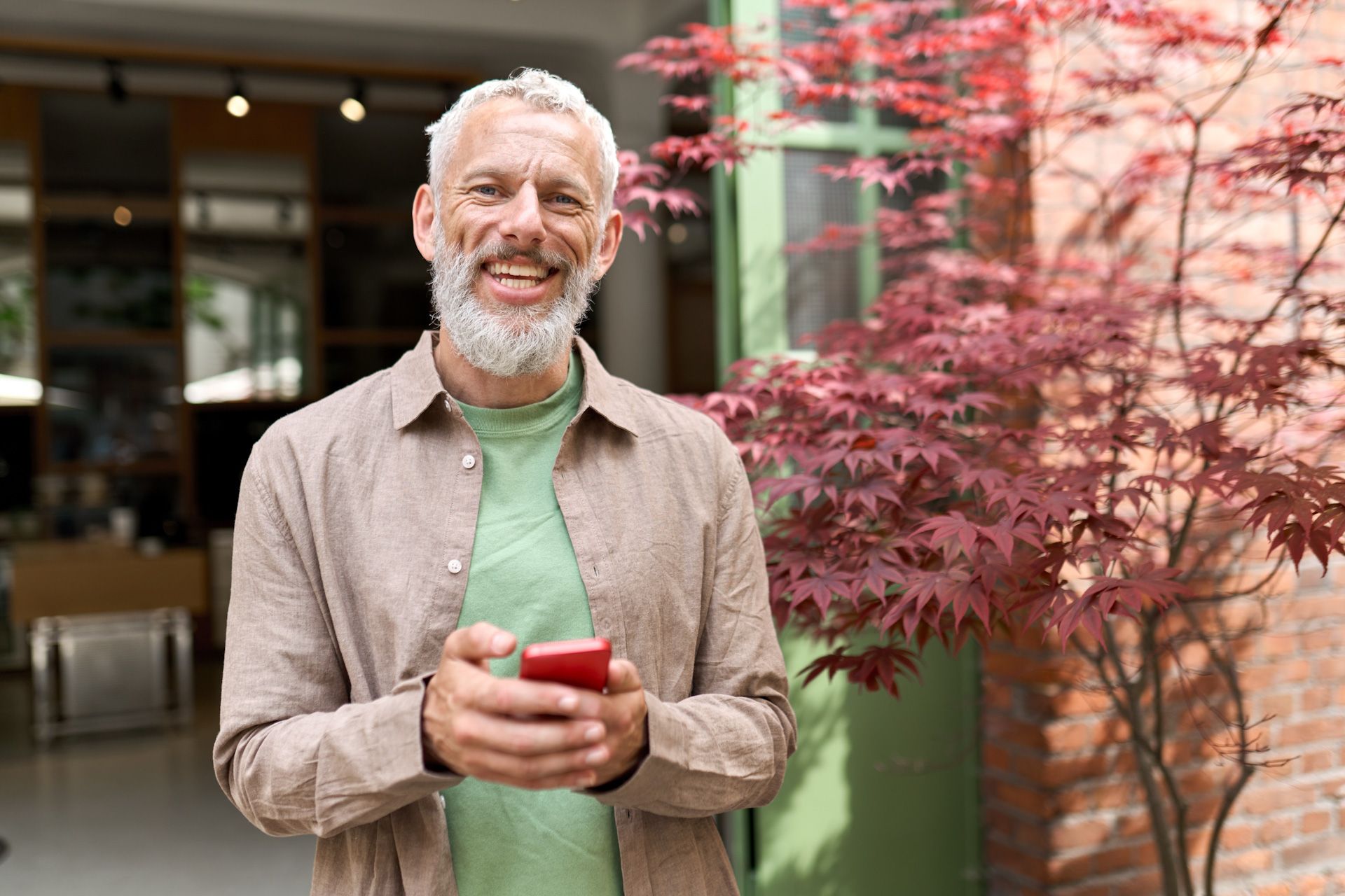 An older man is standing in front of a building holding a cell phone.