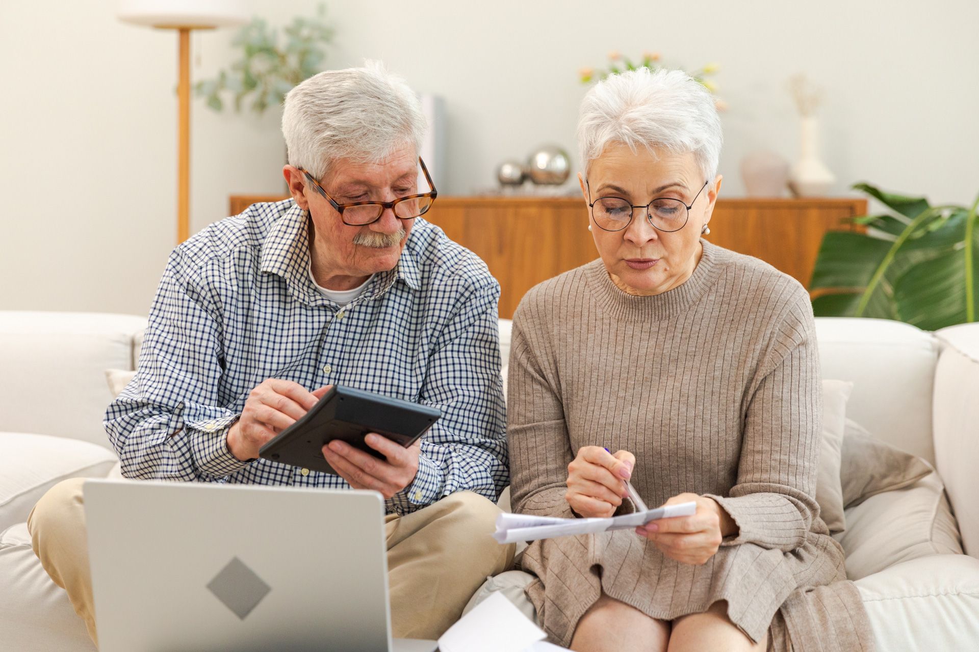 An elderly couple is sitting on a couch looking at a laptop and a tablet.