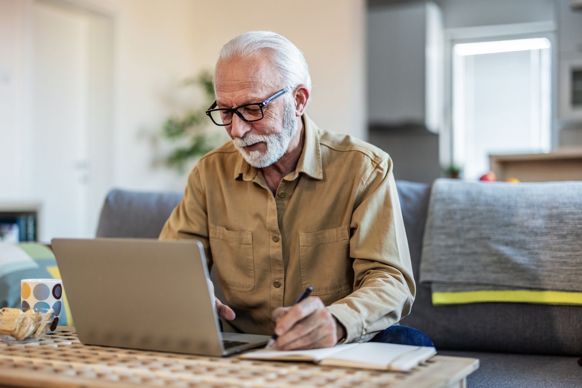 An elderly man is sitting on a couch using a laptop computer.