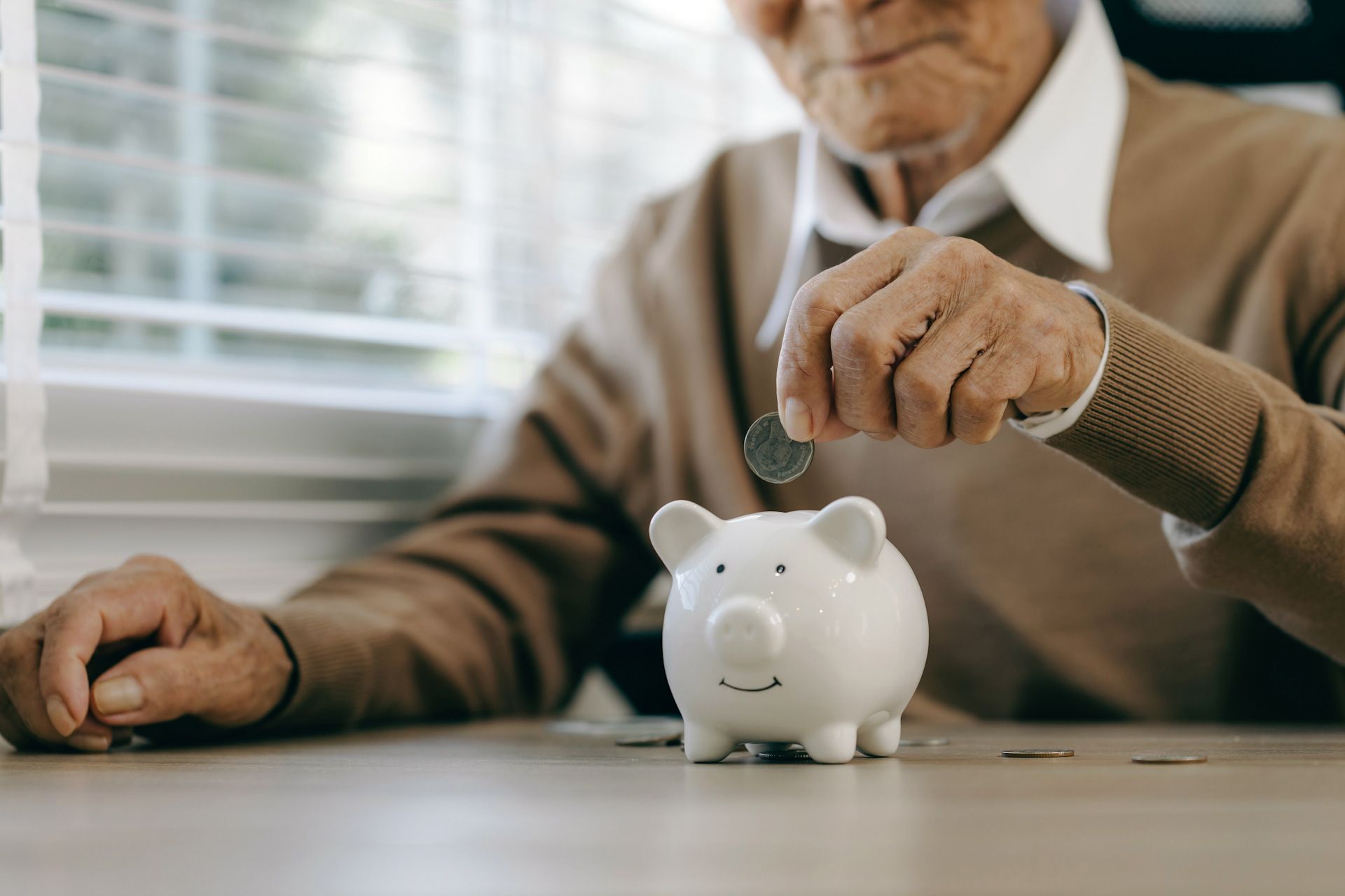 An elderly man is putting a coin into a piggy bank.