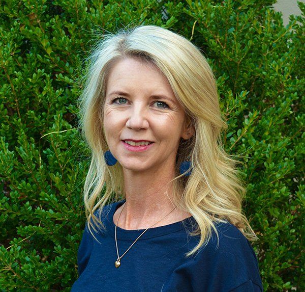 A woman wearing a blue shirt and earrings is standing in front of a bush.