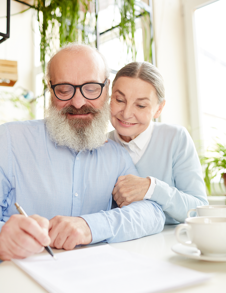 Older man with beard signing document, woman smiles beside him at a table.