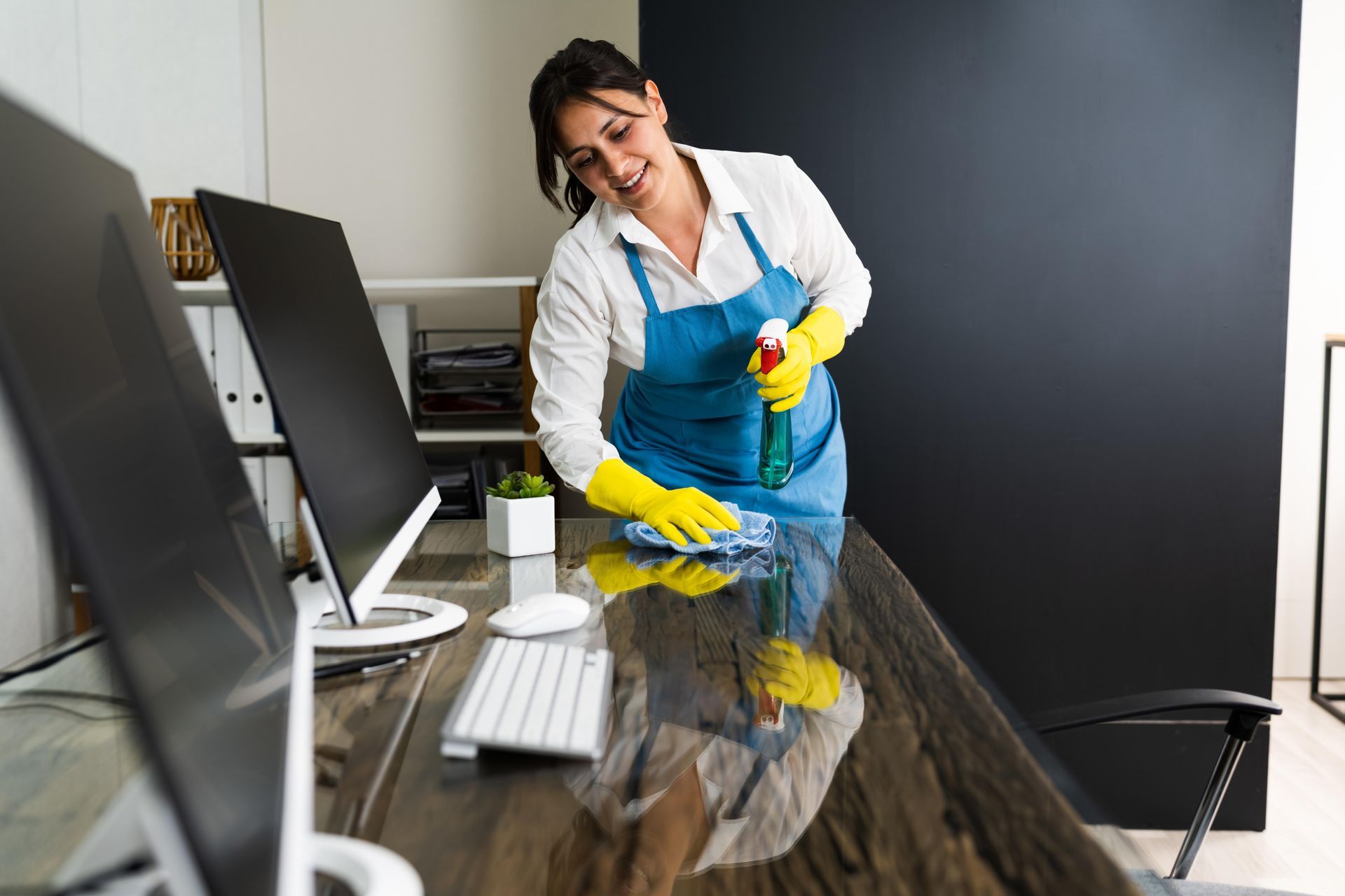 Cleaner in gloves wiping a conference table beside a computer monitor and keyboard in an office
