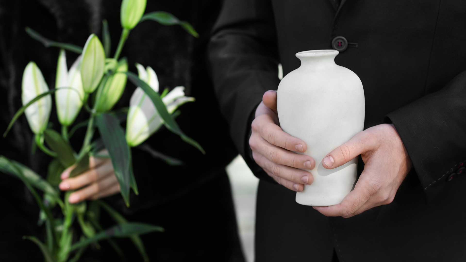 Two people, one holding a white urn, the other lilies, at a funeral.