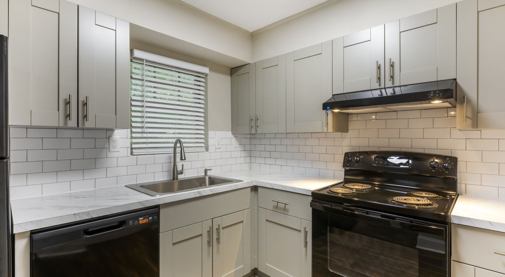 Gray kitchen with white subway tile backsplash, black appliances, and a window with blinds.