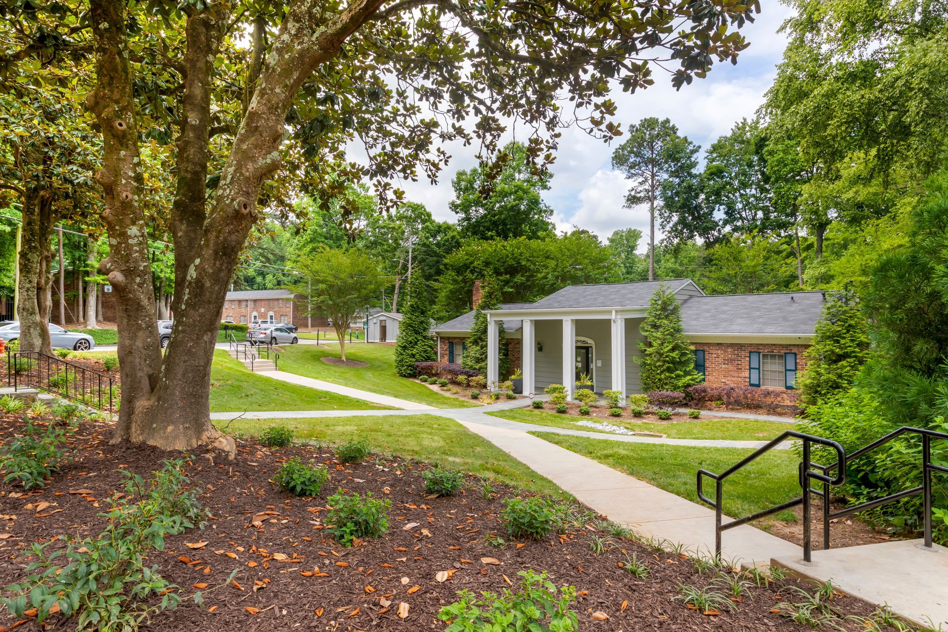 Ranch-style house with brick and gray exterior, pillars, surrounded by greenery and lawn.