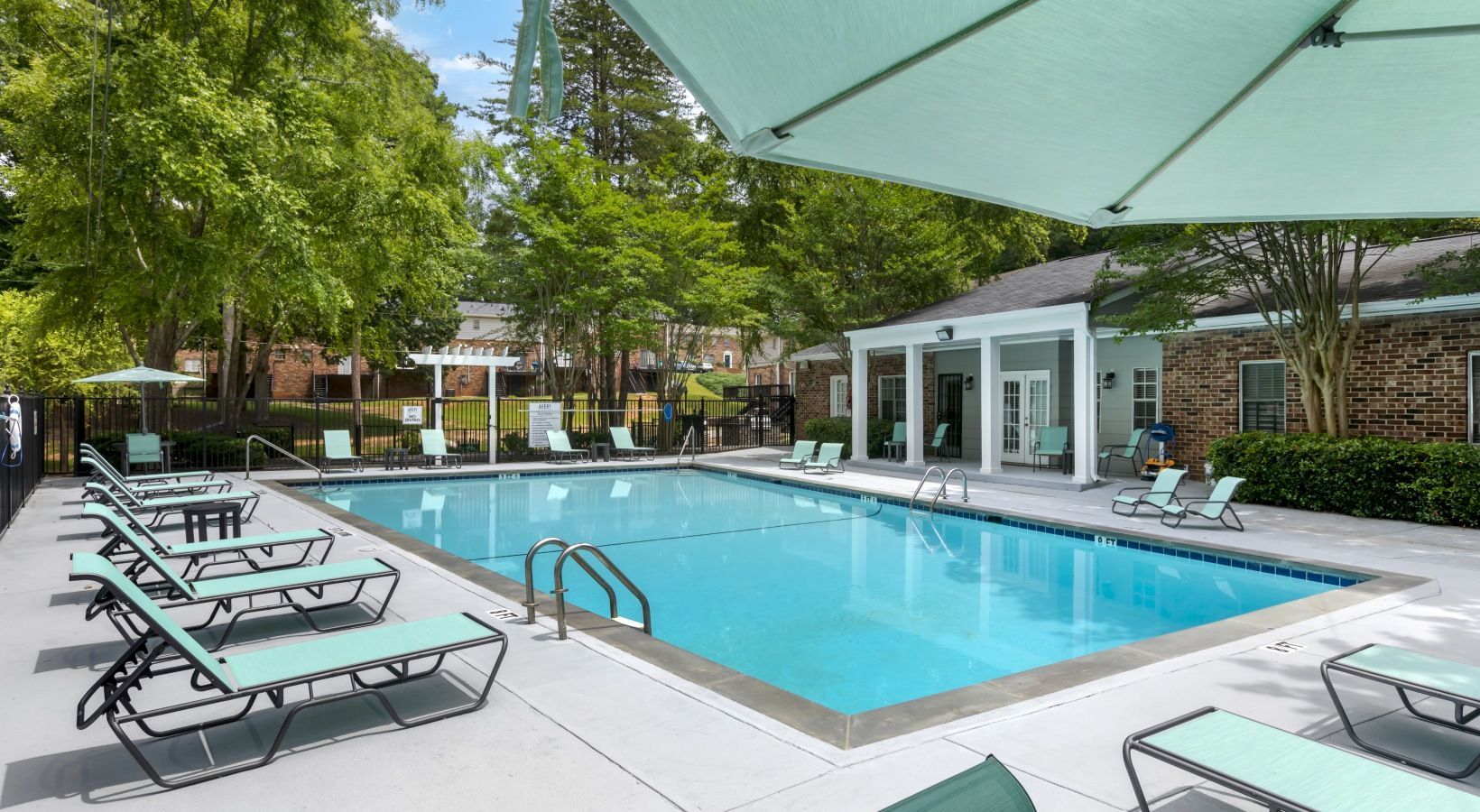 Swimming pool with lounge chairs and parasols, surrounded by trees and a building.