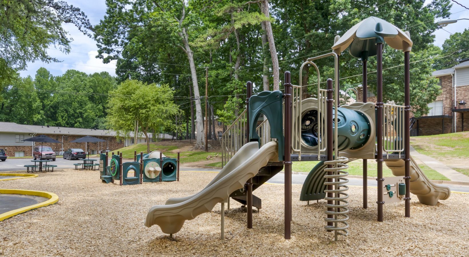 Playground with slides, tunnels, and climbing structures on a wood chip surface, surrounded by trees.
