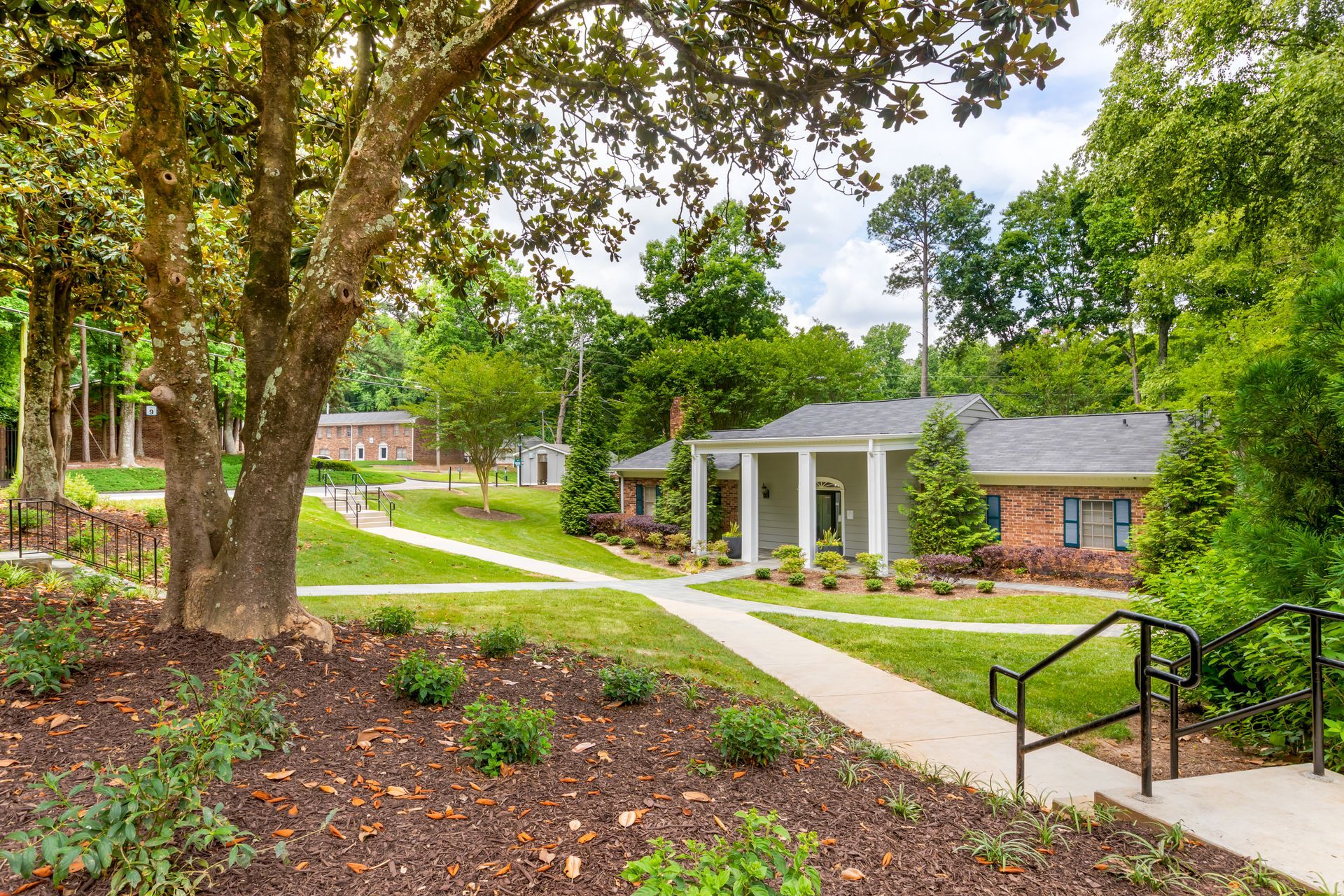 Ranch-style house with brick and gray exterior, pillars, surrounded by greenery and lawn.