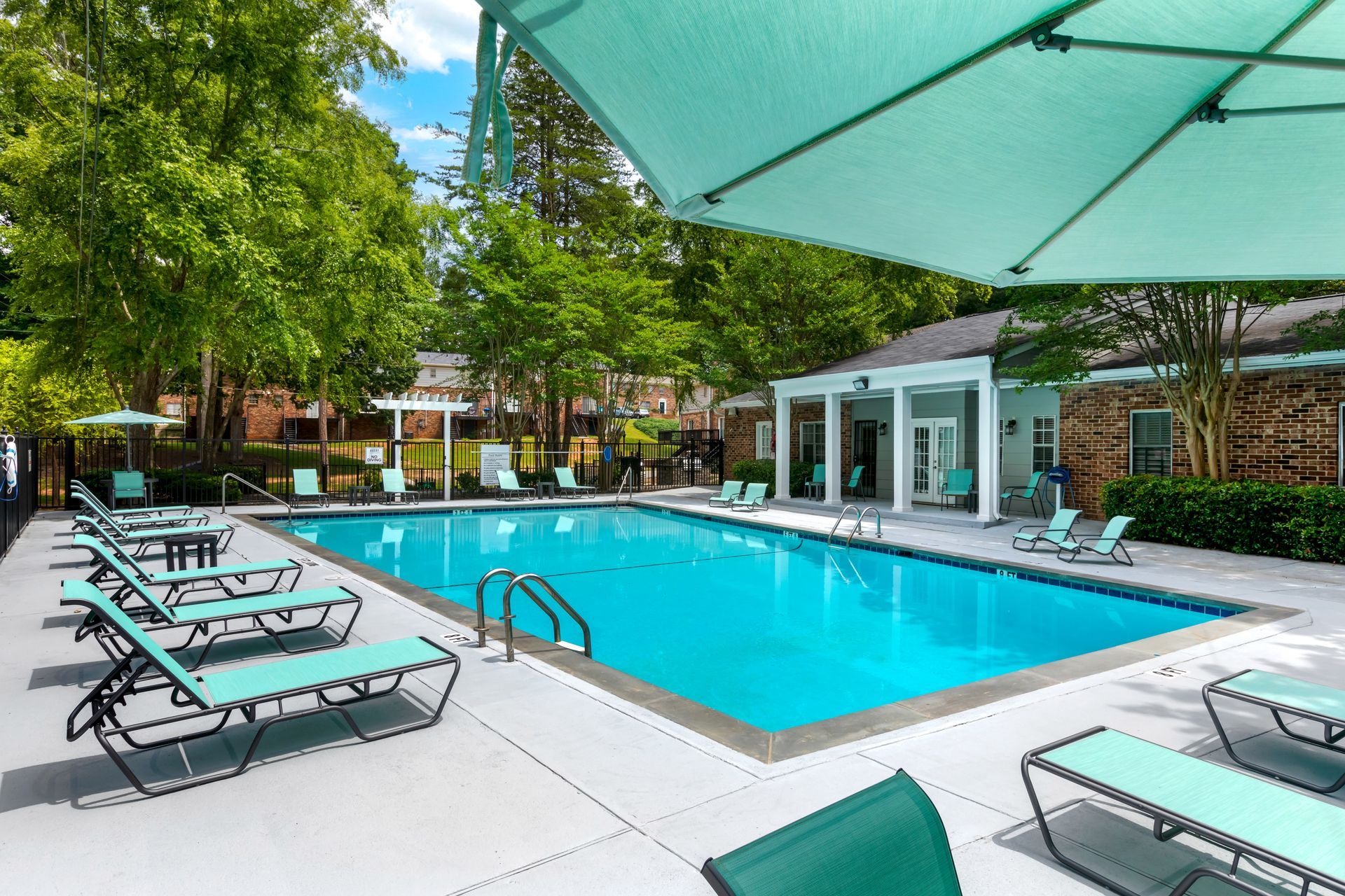 Swimming pool with lounge chairs and parasols, surrounded by trees and a building.