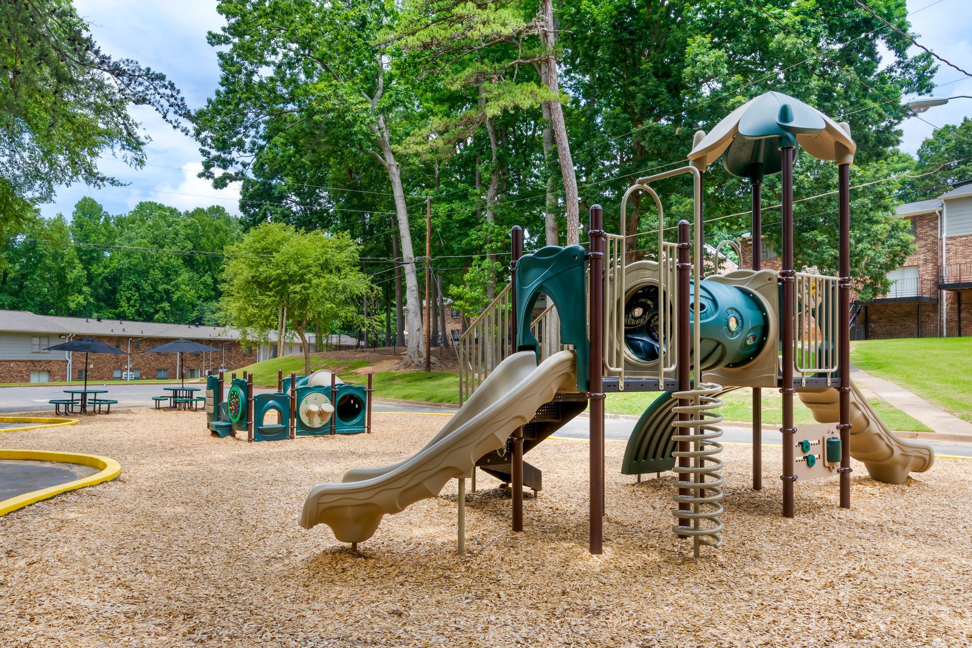 Playground with slides, tunnels, and climbing structures on a wood chip surface, surrounded by trees.