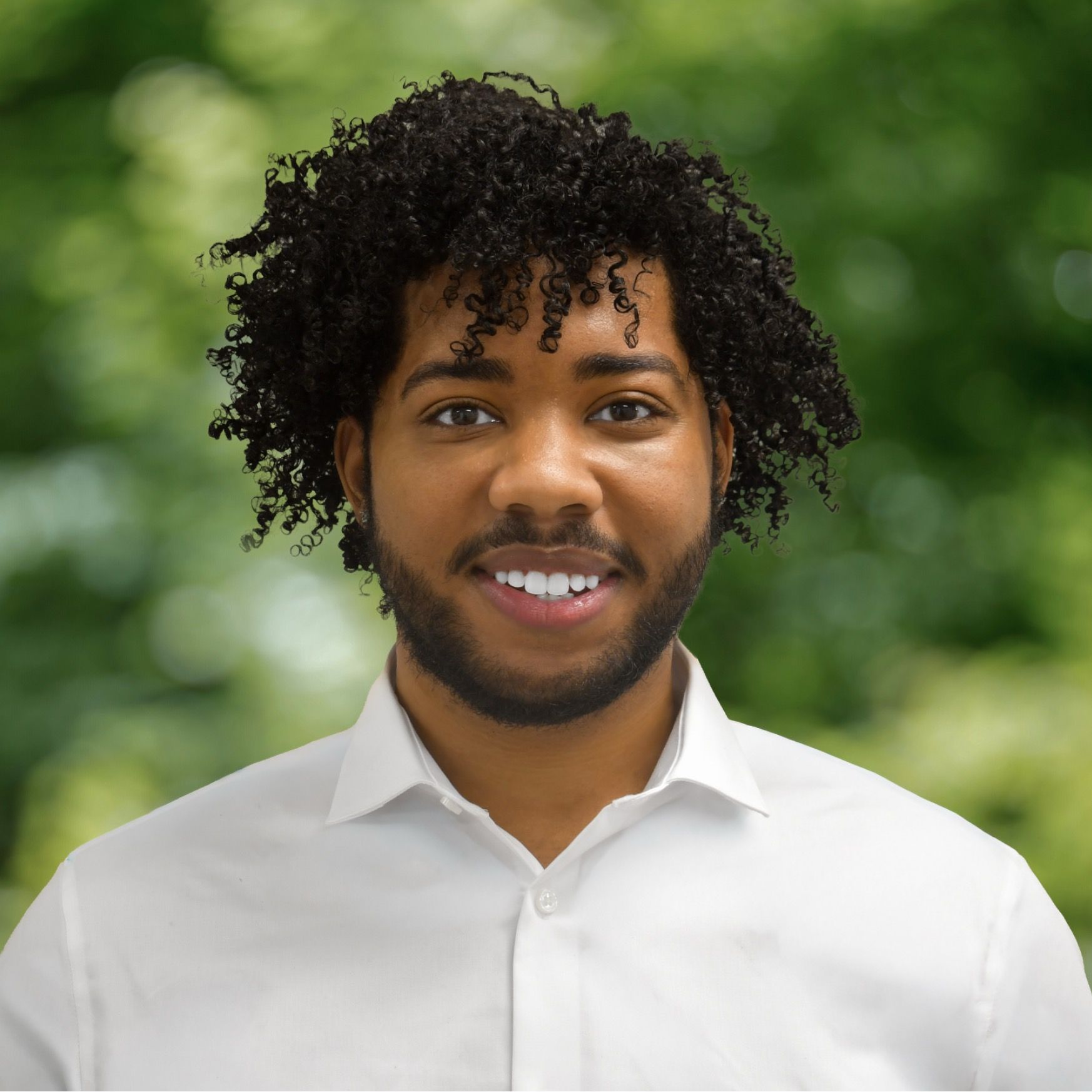 Man with dark hair and beard, wearing striped shirt, smiling, and gazing at the camera, in a bright setting.