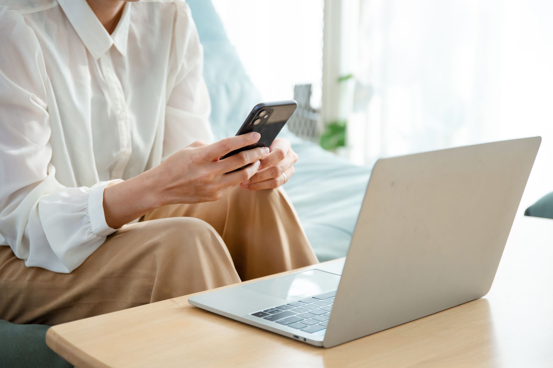 Personne tenant un téléphone, assise, utilisant un ordinateur portable sur une table devant elle, près d'une fenêtre.