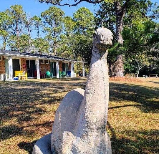 A statue of a dinosaur is sitting in the grass in front of a house.