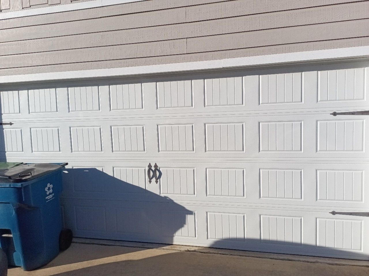 White garage door with decorative panels and black hardware; blue trash can in the foreground.