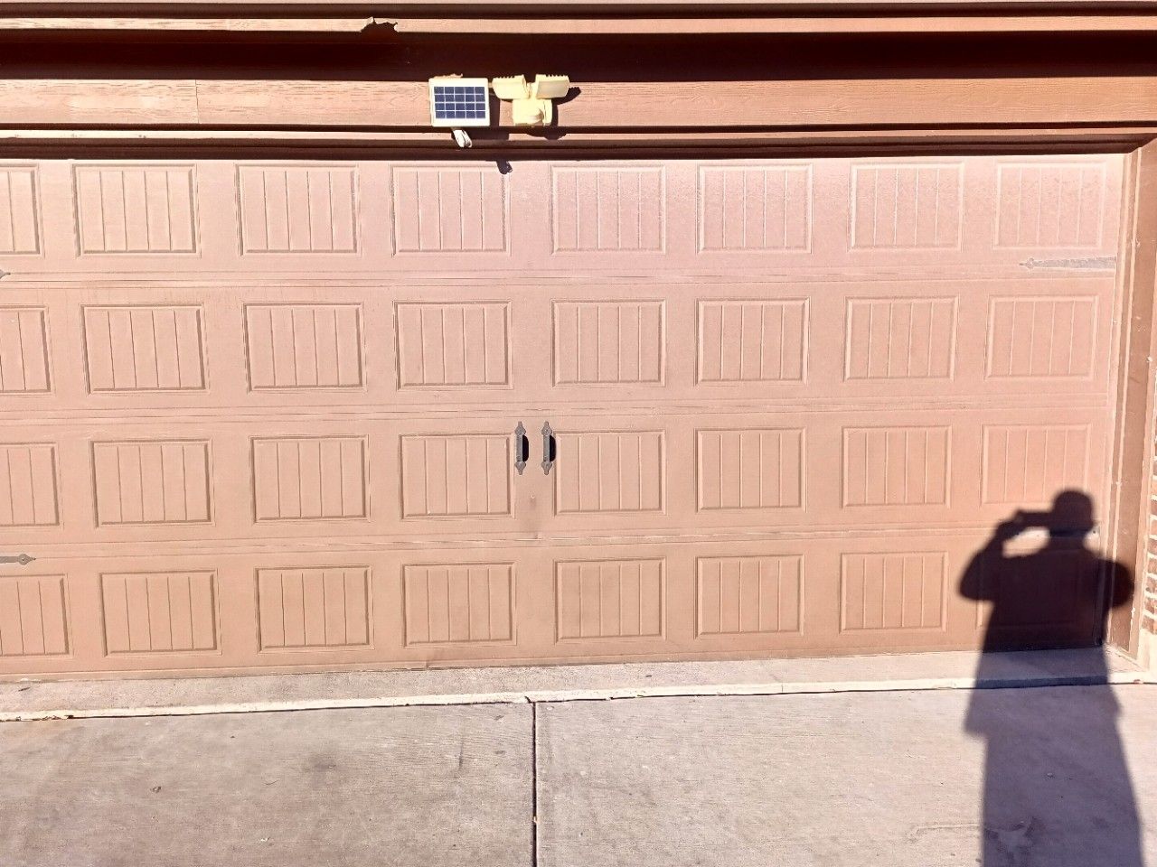 Brown garage door with solar-powered light above. Person's shadow taking photo in sunlight.