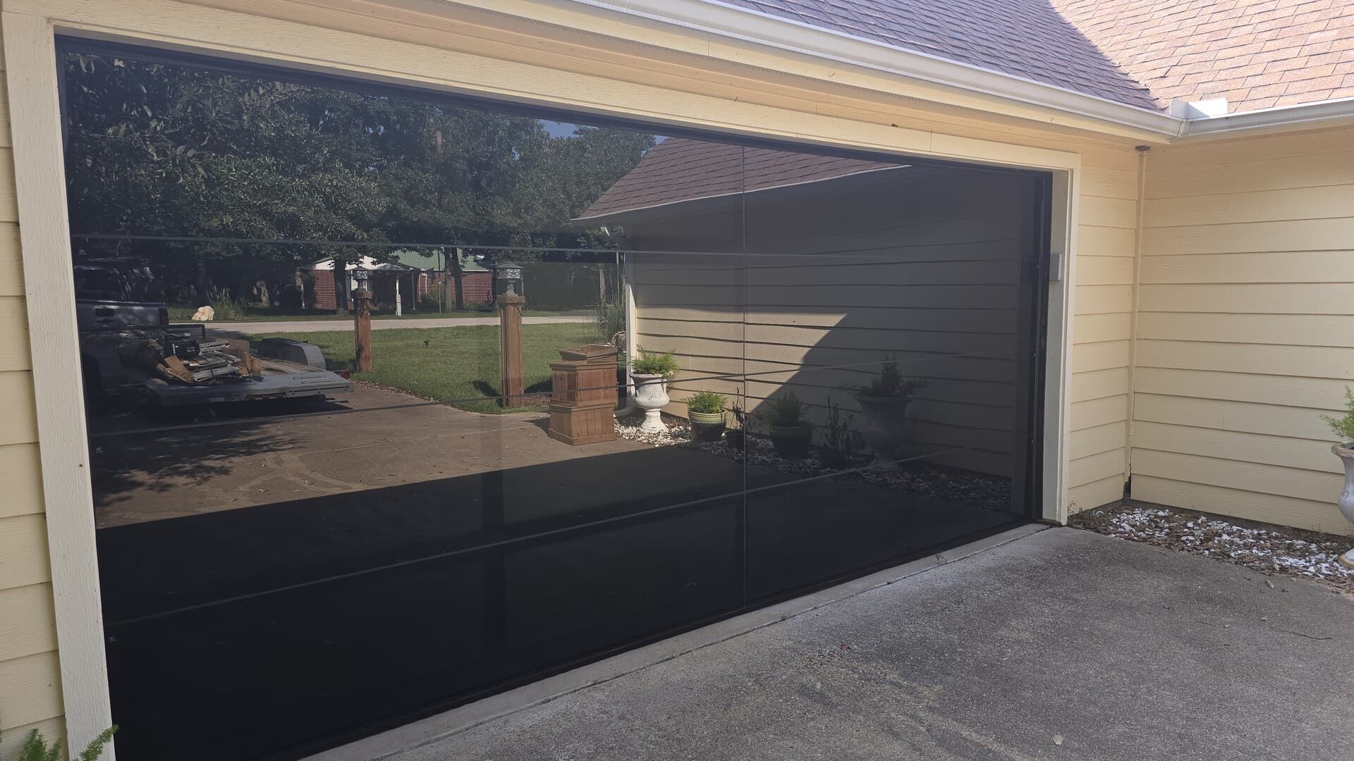 Black garage door reflecting the outdoors; trees, a fence, and yard visible.
