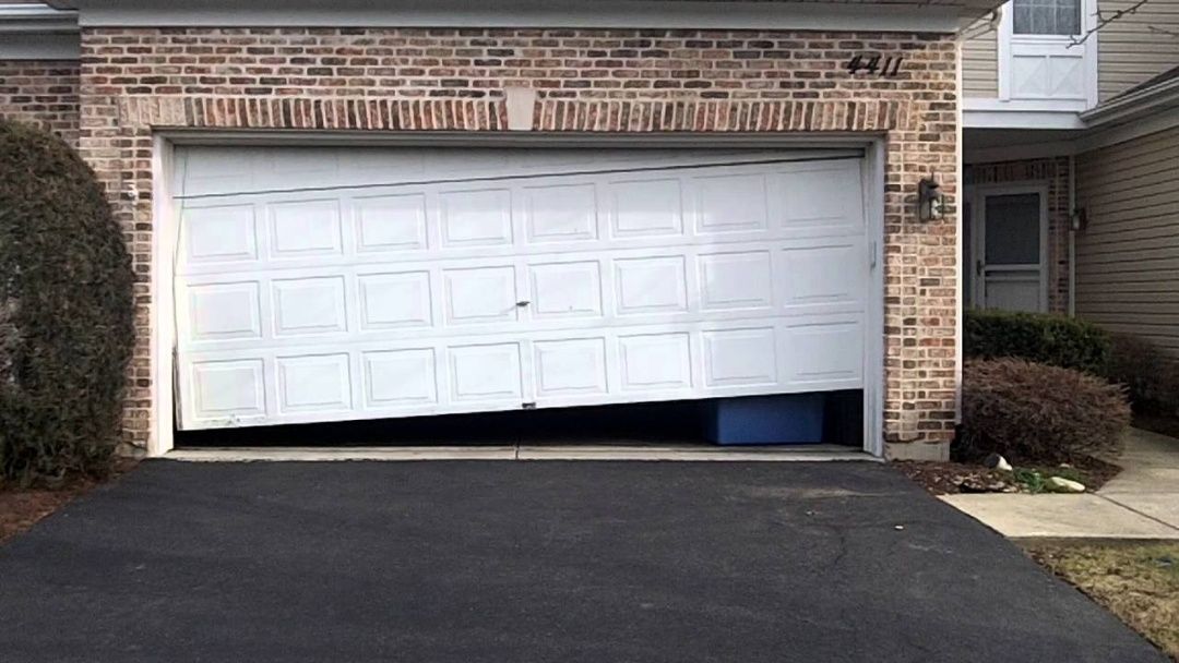 Garage door, partially open and tilted, revealing a blue bin. Brick building exterior.