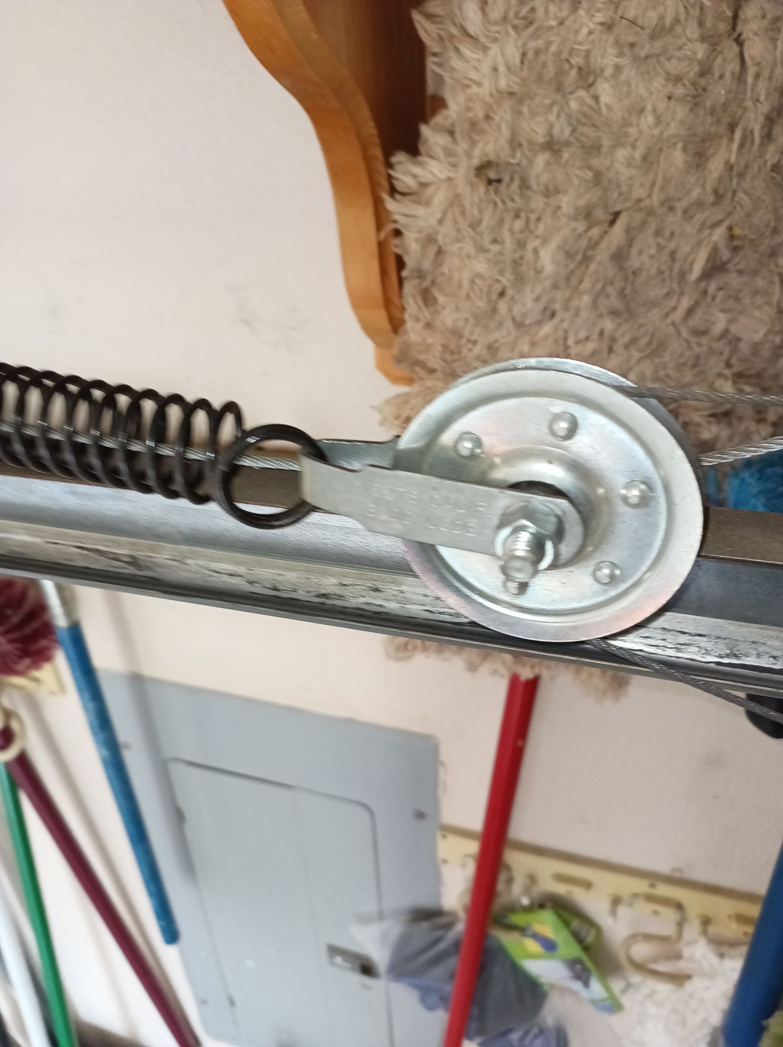 Garage door spring and pulley assembly. Silver spring, pulley, chain, and mounting hardware against a cluttered wall.
