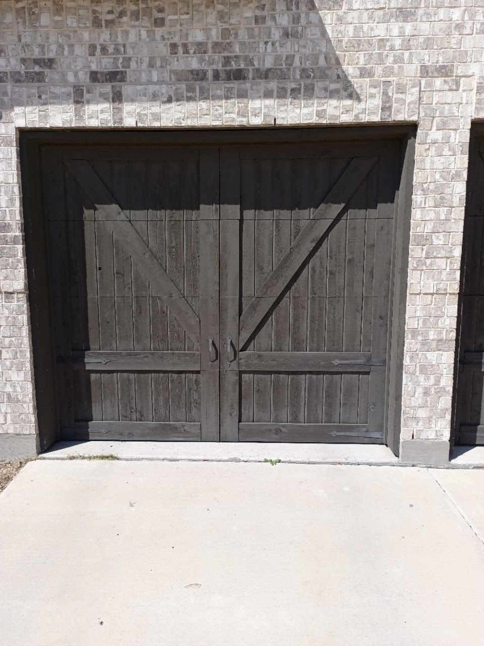 Two gray wooden garage doors with diagonal supports against a brick building, set on a concrete pad.