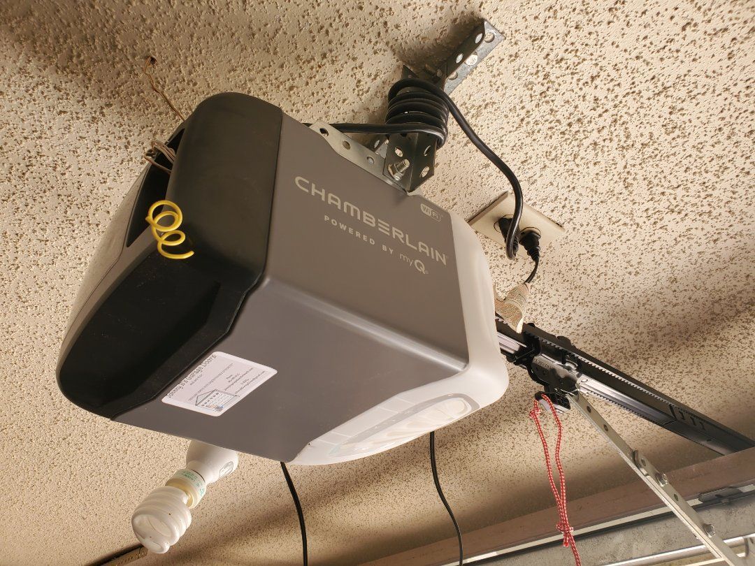 Chamberlain garage door opener mounted on a textured ceiling. Gray and black unit with attached arm and wires.