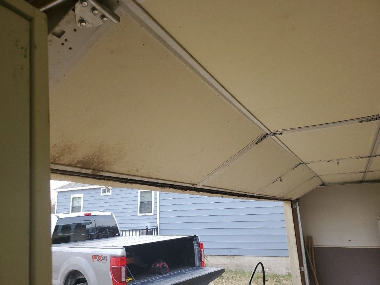 Garage door partially open, revealing a white pickup truck, houses, and the sky. The door is beige.