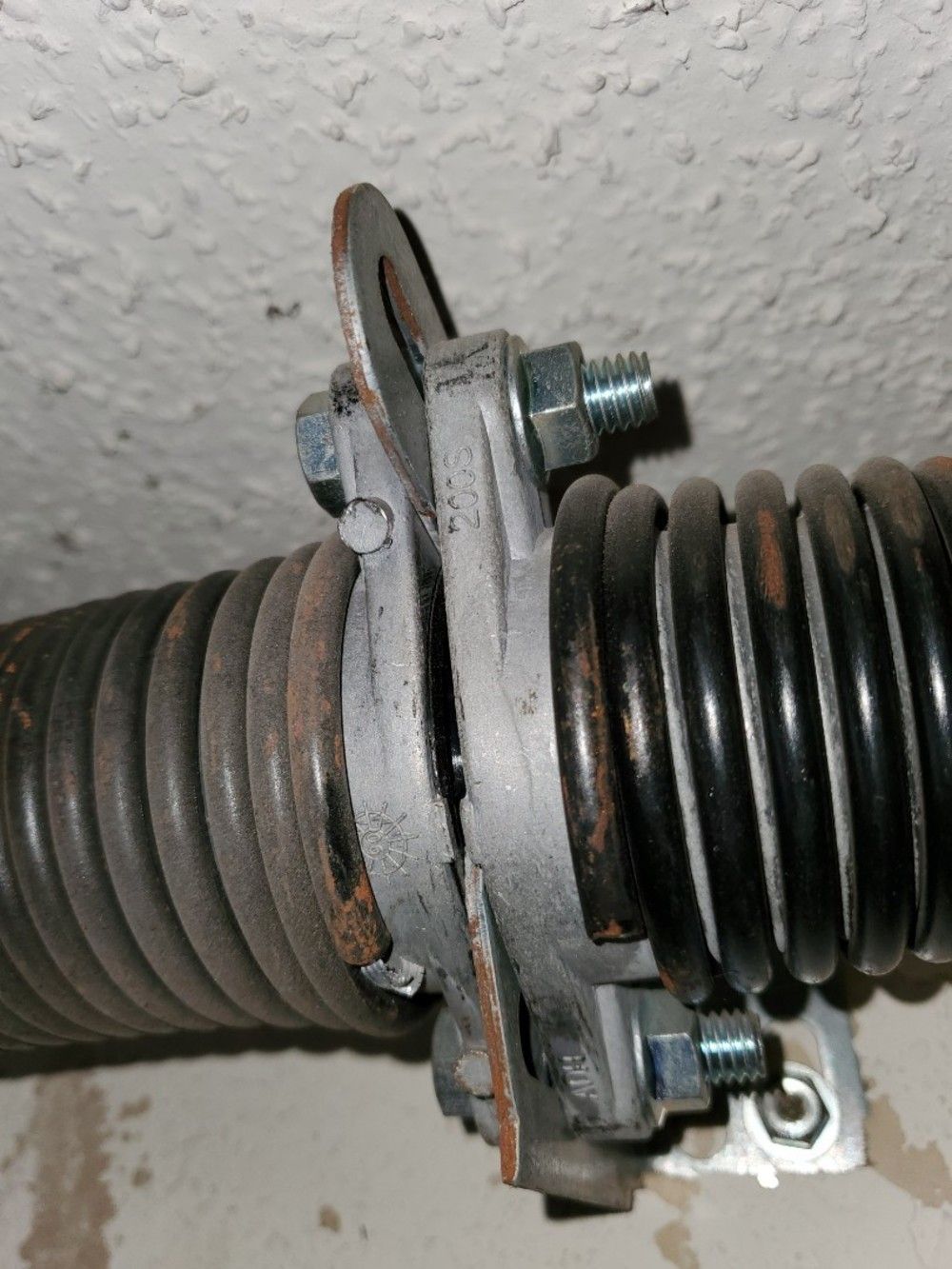 Garage door torsion spring, close-up.  Metal spring is black and gray, attached to a bracket with bolts.