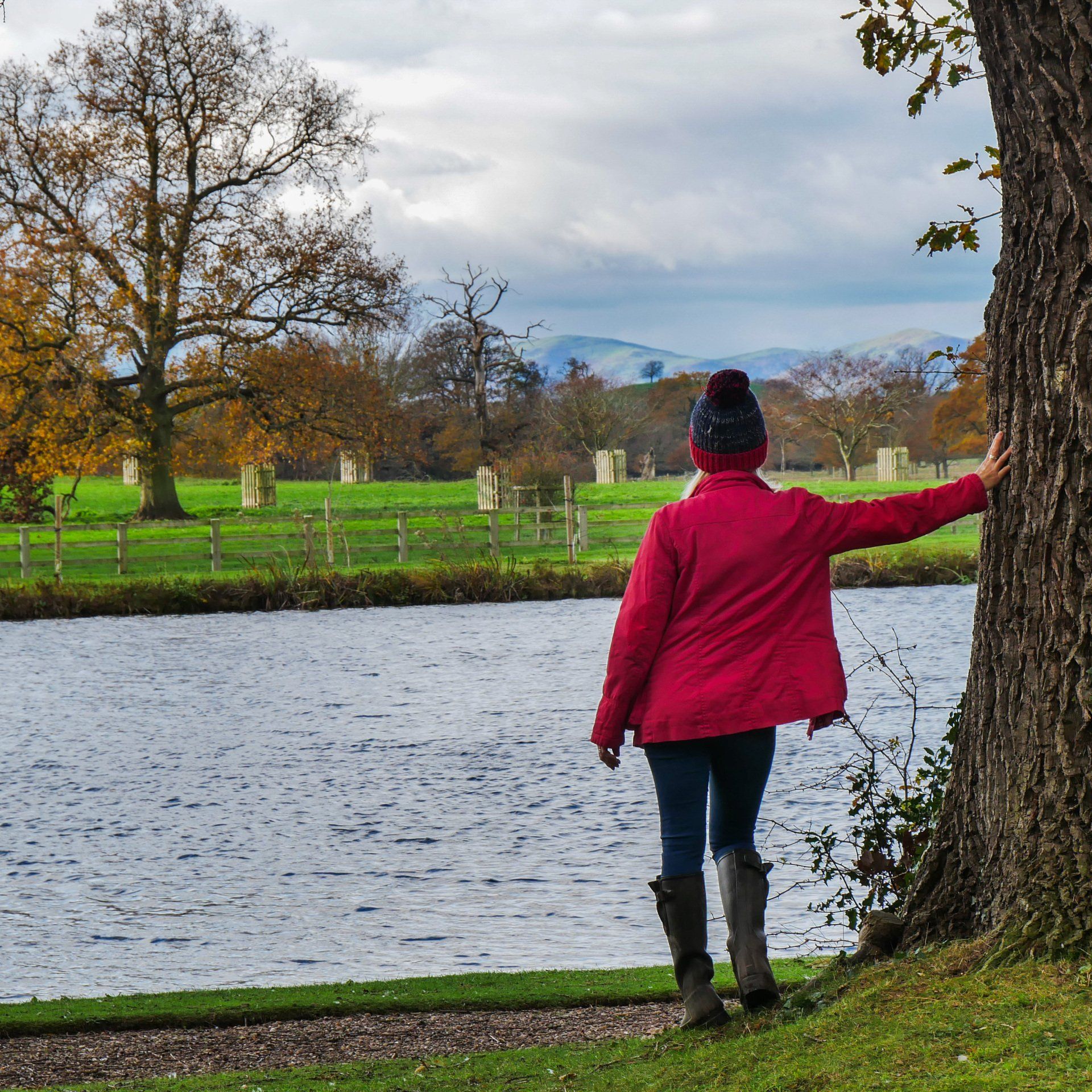 A woman in a red jacket leans against a tree near a body of water
