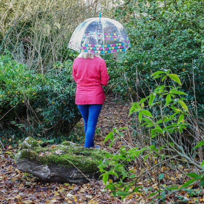 A woman in a pink jacket is walking through the woods with an umbrella.