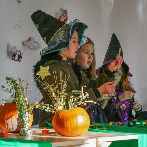 Three young girls dressed as witches are sitting at a table with pumpkins and candles