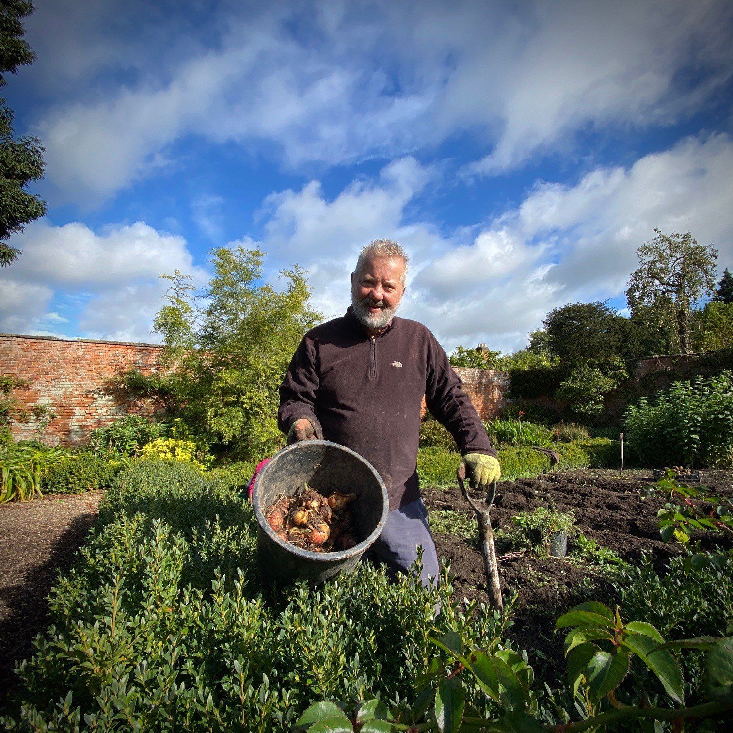 A man is kneeling down in a garden holding a bucket of dirt.