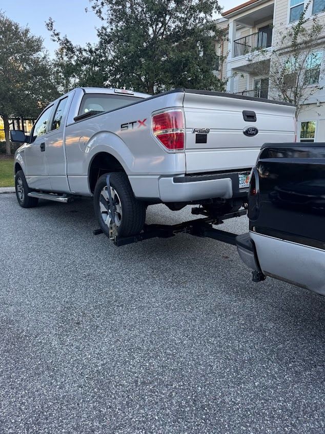 Silver Ford pickup truck towing a black truck in a parking lot.