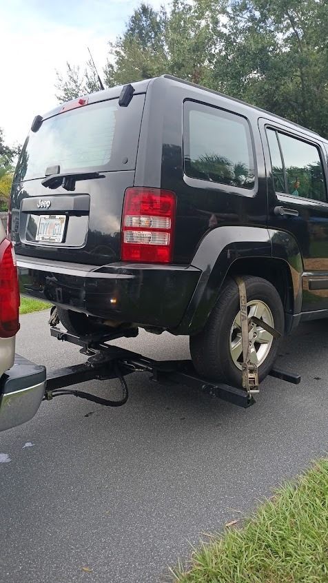Black Jeep Liberty being towed by a truck with its rear wheels on a dolly, on a paved road.