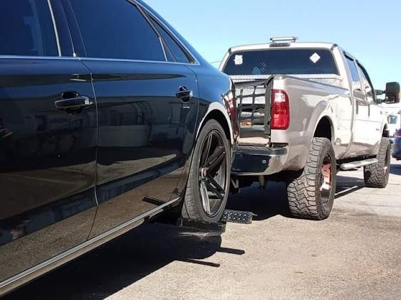 Black car being towed by a tan pickup truck. The car's flat tire rests on a loading ramp. Sunny day.