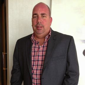 Man in dark suit jacket and plaid shirt stands indoors, facing the camera.