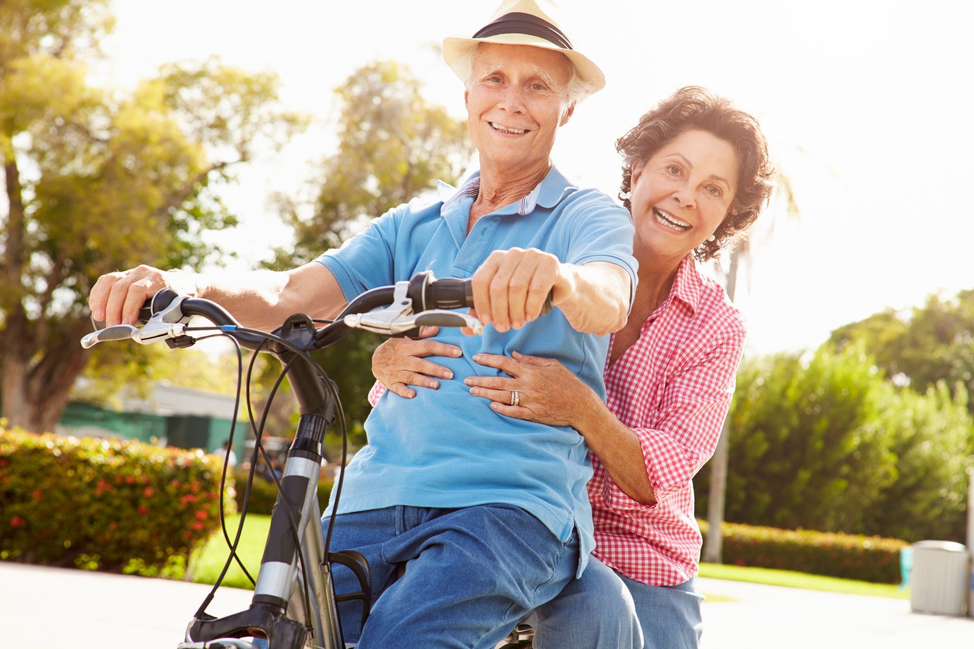 Smiling couple on a bicycle outdoors; woman embraces man, both look at camera.